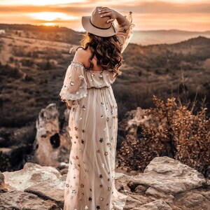 May include: A beige off-the-shoulder dress with sheer sleeves and a flowing skirt, adorned with star-shaped sequins. A wide-brimmed hat and long, wavy hair complete the look. The dress is worn outdoors with a scenic mountain backdrop at sunset.
