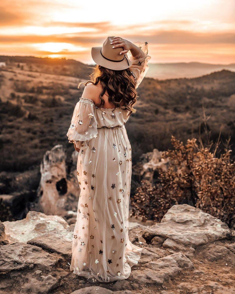 May include: A beige off-the-shoulder dress with sheer sleeves and a flowing skirt, adorned with star-shaped sequins. A wide-brimmed hat and long, wavy hair complete the look. The dress is worn outdoors with a scenic mountain backdrop at sunset.