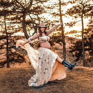 May include: A pregnant person wearing a beige crop top and a long, flowing skirt adorned with star-shaped embellishments. The outfit is paired with a brown hat and black cowboy boots. The photo is taken outdoors with a backdrop of trees.