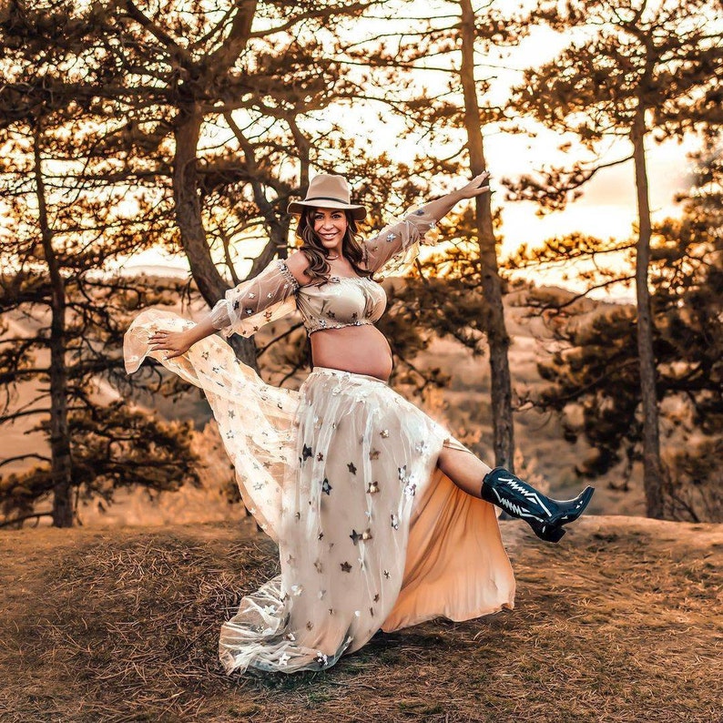May include: A pregnant person wearing a beige crop top and a long, flowing skirt adorned with star-shaped embellishments. The outfit is paired with a brown hat and black cowboy boots. The photo is taken outdoors with a backdrop of trees.