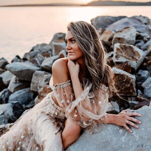 May include: A beige two-piece outfit with star embellishments. The top has off-the-shoulder sleeves with a sheer overlay, and the skirt is flowing. The outfit is worn by a person sitting on rocks near a body of water at sunset.