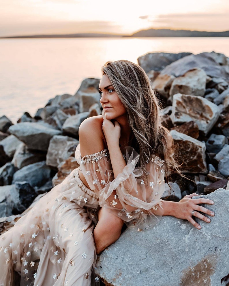 May include: A beige two-piece outfit with star embellishments. The top has off-the-shoulder sleeves with a sheer overlay, and the skirt is flowing. The outfit is worn by a person sitting on rocks near a body of water at sunset.