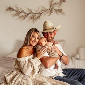 May include: A family portrait featuring a mother, father, and newborn baby. The mother wears an off-the-shoulder dress, and the father wears a cowboy hat and glasses. The baby is wrapped in a blanket. The background includes a decorative wall hanging.