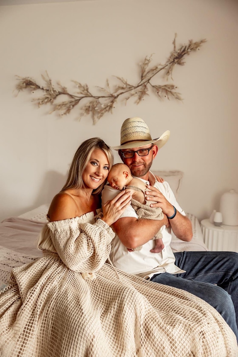 May include: A family portrait featuring a mother, father, and newborn baby. The mother wears an off-the-shoulder dress, and the father wears a cowboy hat and glasses. The baby is wrapped in a blanket. The background includes a decorative wall hanging.