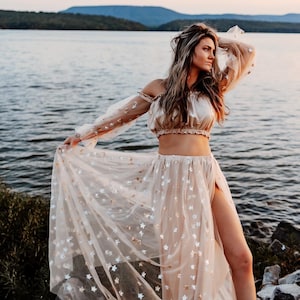 May include: A beige two-piece outfit with a crop top and a long skirt. The skirt has a high slit and is adorned with silver star-shaped sequins. The top has off-the-shoulder sleeves. The outfit is worn outdoors near a lake.