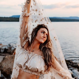 May include: A beige two-piece outfit with star embellishments. The top is a cropped, off-the-shoulder style. The sheer, long-sleeved overlay is adorned with gold and silver stars. The outfit is worn near a body of water.