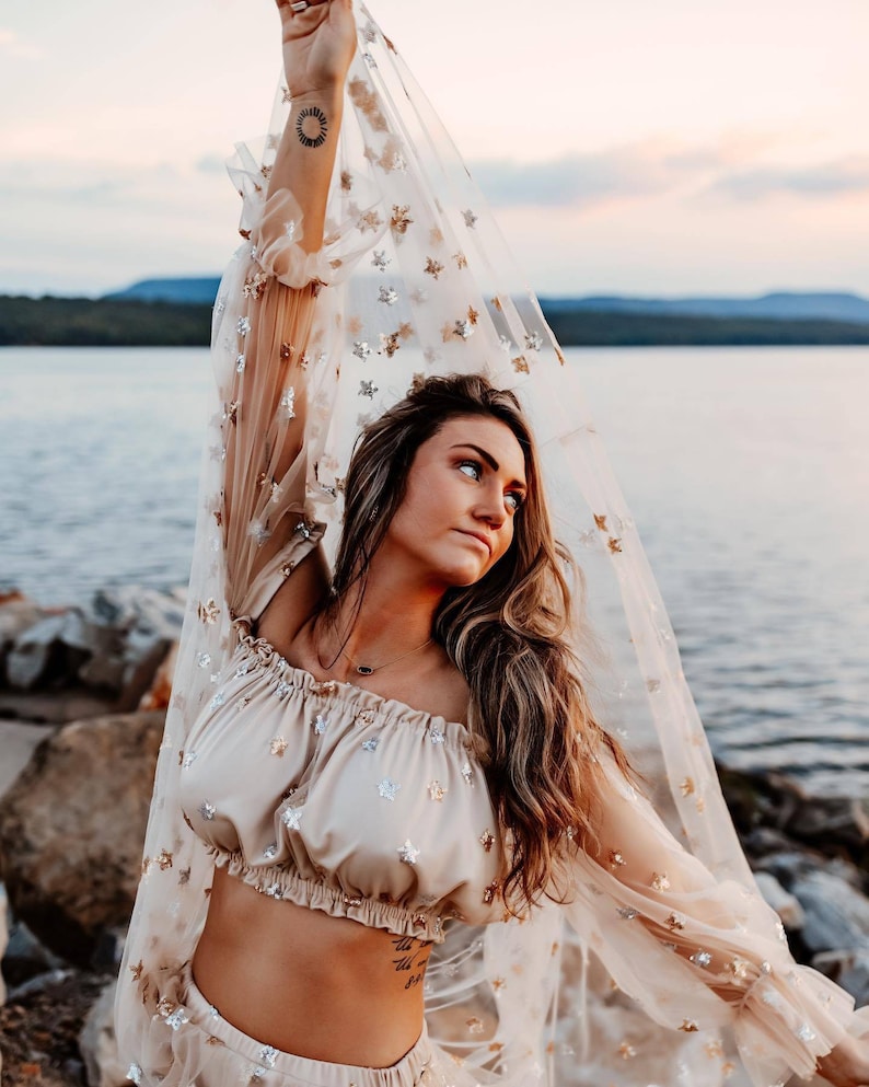 May include: A beige two-piece outfit with star embellishments. The top is a cropped, off-the-shoulder style. The sheer, long-sleeved overlay is adorned with gold and silver stars. The outfit is worn near a body of water.