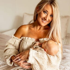 May include: A woman with long blonde hair smiles while holding a baby. She wears an off-the-shoulder cream-colored top with a textured, open-weave design. The baby is wrapped in a matching blanket. The background is a soft, neutral color.