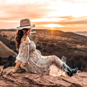 May include: A beige hat with a decorative band sits atop a person's head. The person is wearing a sheer, off-the-shoulder dress adorned with star-shaped embellishments and black cowboy boots. The background features a sunset over a mountainous landscape.