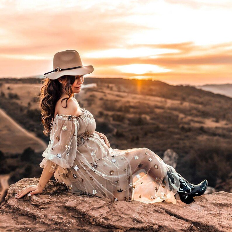 May include: A beige hat with a decorative band sits atop a person's head. The person is wearing a sheer, off-the-shoulder dress adorned with star-shaped embellishments and black cowboy boots. The background features a sunset over a mountainous landscape.