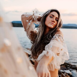 May include: A beige off-the-shoulder top with sheer, ruffled sleeves adorned with silver star-shaped sequins. The model is standing near a body of water, with long, light brown hair.