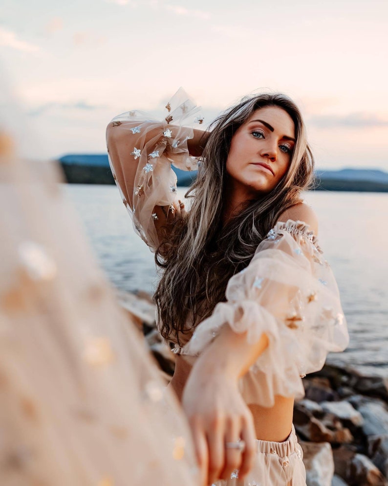 May include: A beige off-the-shoulder top with sheer, ruffled sleeves adorned with silver star-shaped sequins. The model is standing near a body of water, with long, light brown hair.