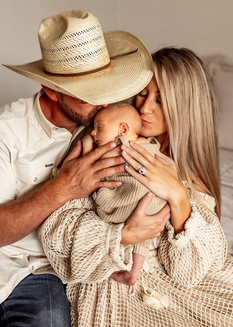 May include: A family portrait featuring a newborn baby held by parents. The father wears a straw cowboy hat and a button-down shirt. The mother has long blonde hair and a ring on her finger. The baby is wrapped in a beige knit blanket.