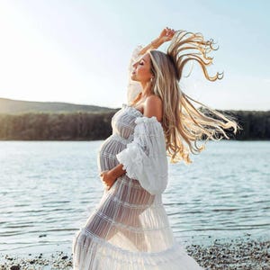 May include: A white, off-the-shoulder maternity dress with long, ruffled sleeves. The dress is sheer, revealing the silhouette of a pregnant person. The person's long, blonde hair is flowing in the wind, and they are standing near a body of water.