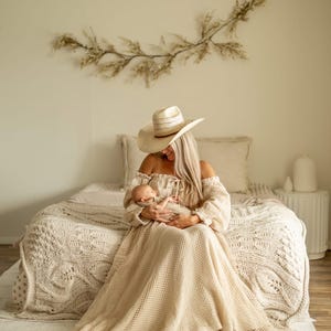 May include: A woman wearing a beige off-the-shoulder dress and a straw hat, holding a newborn baby. The dress has long sleeves and a flowing skirt. The room has a neutral color palette with a decorative branch on the wall.