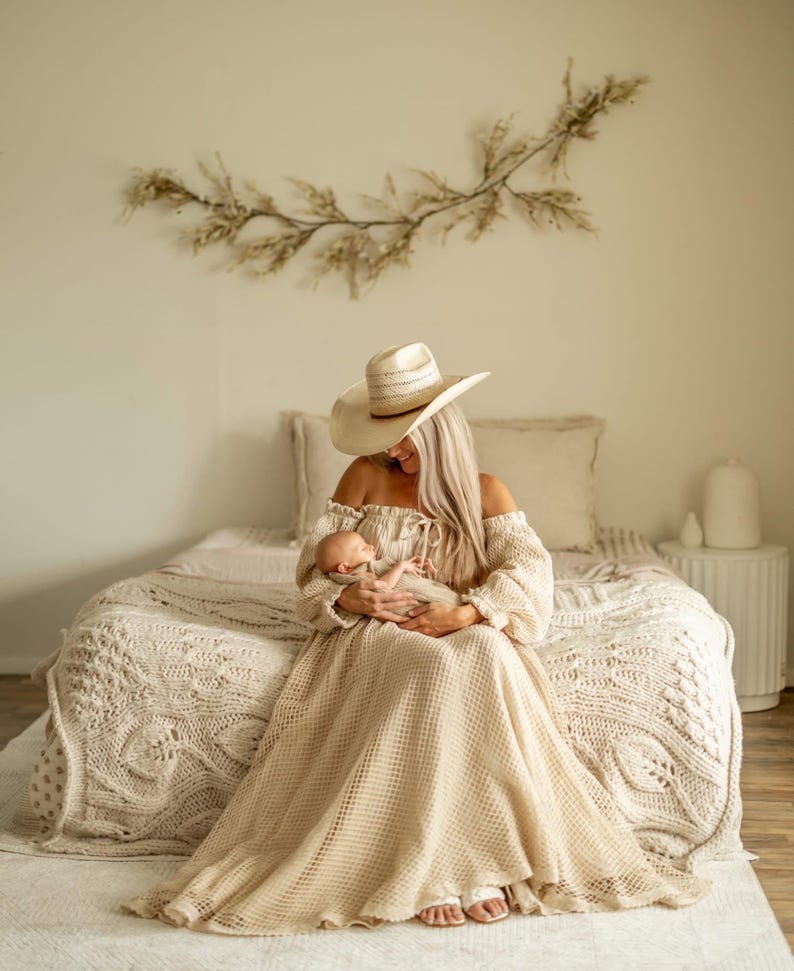 May include: A woman wearing a beige off-the-shoulder dress and a straw hat, holding a newborn baby. The dress has long sleeves and a flowing skirt. The room has a neutral color palette with a decorative branch on the wall.