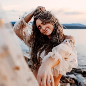 May include: A woman wearing a beige off-the-shoulder top with sheer, ruffled sleeves adorned with silver embellishments. Her long, wavy hair is styled down, and she is smiling with her eyes closed. The background features a body of water and a soft, pastel sky.