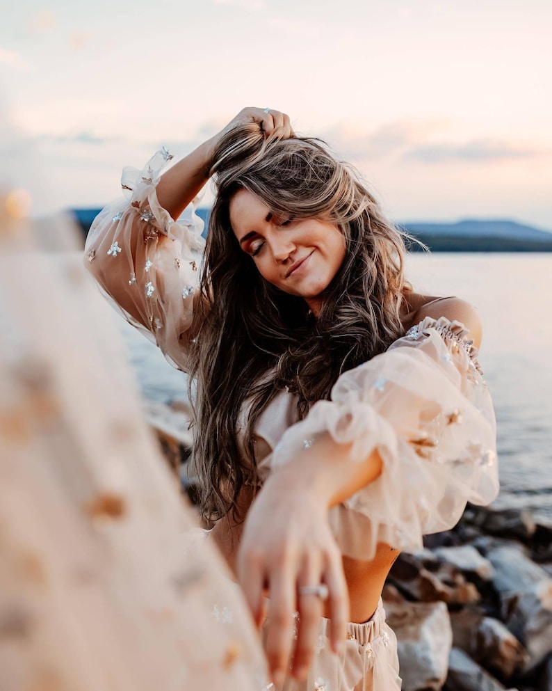 May include: A woman wearing a beige off-the-shoulder top with sheer, ruffled sleeves adorned with silver embellishments. Her long, wavy hair is styled down, and she is smiling with her eyes closed. The background features a body of water and a soft, pastel sky.