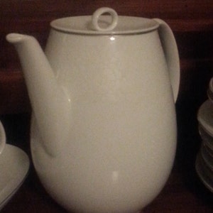 May include: White ceramic teapot with a lid and a gold trim around the top. The teapot has a rounded body and a spout on the side.