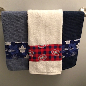 May include: Three towels hanging on a towel rack. The towel on the left is blue with a Toronto Maple Leafs design. The towel in the middle is white with a red and black plaid design. The towel on the right is navy blue with a Toronto Maple Leafs design.