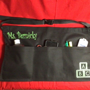 May include: Black apron with multiple pockets, featuring the embroidered name "Ms. Bernicky" in green. The apron has a black strap and buckle. The apron contains a bottle, phone, pens, and a tag. The lower right corner has a green ABC block design.