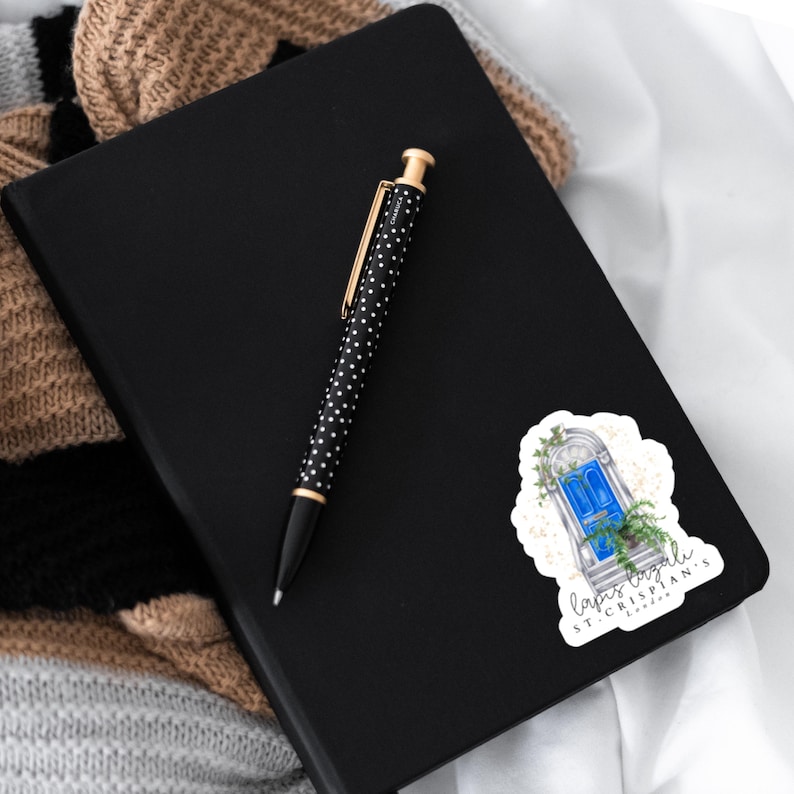 Puede incluir: Un cuaderno negro con un bol&iacute;grafo de lunares blancos y negros y una pegatina con una puerta azul con el texto "Lapis Lazuli St. Crispian's London". El bol&iacute;grafo tiene detalles dorados.