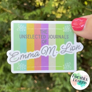 May include: A sticker with the text "The Unselected Journals of Emma M. Lion" in a decorative font. The sticker has a striped background in pastel colors and floral accents. The sticker also has the text "NorahLu Designs".