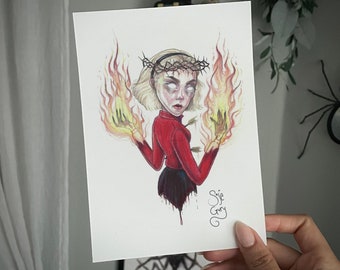 Puede incluir: Una ilustración dibujada a mano de una mujer con cabello rubio que lleva una corona de espinas y un vestido rojo. Ella sostiene llamas en cada mano. La ilustración está firmada "Sage Gary".