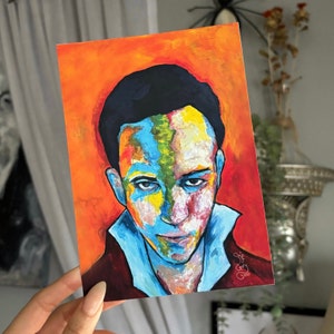 May include: A colorful painting of a person's face. The background is a vibrant orange. The person's face is painted in shades of blue, green, yellow, and white. The painting is signed "Sjaf G" and dated "2022".