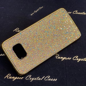 Puede incluir: Una funda de teléfono dorada cubierta de diamantes de imitación iridiscentes. La funda tiene un recorte para el objetivo de la cámara. La funda está sobre un fondo negro con el texto "Rangsee Crystal Cases" impreso en ella.