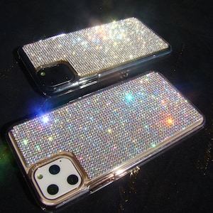 以下が含まれることがあります： 虹色のラインストーンで覆われた2つの金色のiPhoneケース。ケースはカメラの盛り上がりが高く、金色の縁取りがされています。