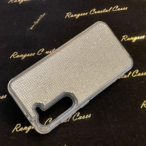 May include: A clear phone case with a sparkly, diamond-like design. The case is made of a clear plastic material and has a raised edge around the camera lens. The case is covered in small, white rhinestones.
