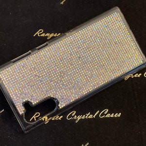 Puede incluir: Una funda transparente para teléfono cubierta de piedras de cristal iridiscentes. La funda está diseñada para un teléfono Samsung Galaxy y tiene un recorte para el objetivo de la cámara. La funda está sobre una superficie de terciopelo negro.