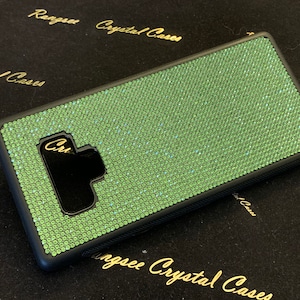 May include: A green phone case covered in sparkling rhinestones. The case has a black border and a cutout for the camera lens.