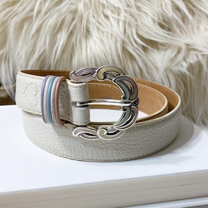 Könnte beinhalten: Ein cremefarbener Gürtel mit einer dekorativen silbernen Schnalle. Der Gürtel hat eine strukturierte Oberfläche und einen mehrfarbigen Akzent in der Nähe der Schnalle. Die Schnalle hat ein einzigartiges, wirbelndes Design. Der Gürtel liegt auf einer weißen Oberfläche.