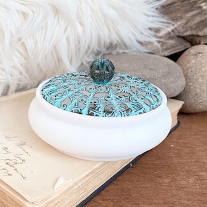 Peut inclure: Une boîte à bijoux en céramique blanche avec un couvercle en filigrane turquoise et un bouton décoratif. La boîte est posée sur un livre ancien ouvert avec du texte manuscrit. L'arrière-plan comprend un tissu blanc duveteux, des pierres et une surface en bois.