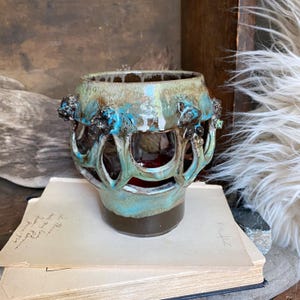 May include: A handcrafted ceramic vase with a unique, openwork design. The vase features a turquoise and brown glaze with dark accents. The interior is a deep red, and the vase sits on a stack of vintage papers.