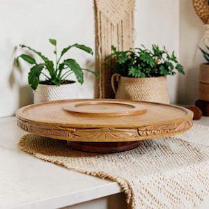 Puede incluir: Un soporte de pedestal redondo de madera de color marrón claro con una superficie superior lisa y plana, y un patrón tallado y texturizado a lo largo del borde exterior. El soporte se apoya en una base cilíndrica central de madera.