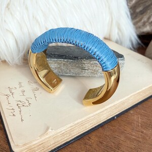Könnte beinhalten: Ein goldfarbenes Armreif-Armband mit einem gewebten hellblauen Band. Das Armband hat ein geschwungenes Design mit poliertem Finish. Das Armband liegt auf einem offenen Buch mit handschriftlichem Text.