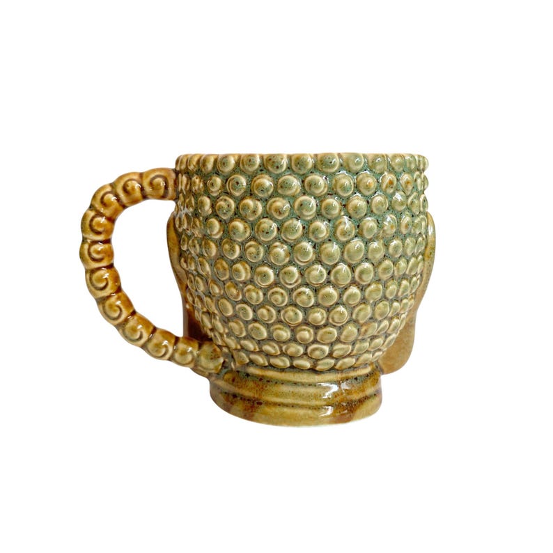 May include: A ceramic mug shaped like a head with a textured surface of small raised bumps in shades of sage green and brown. The handle features a repeating spiral pattern, and the base is a smooth, rounded brown ceramic finish.