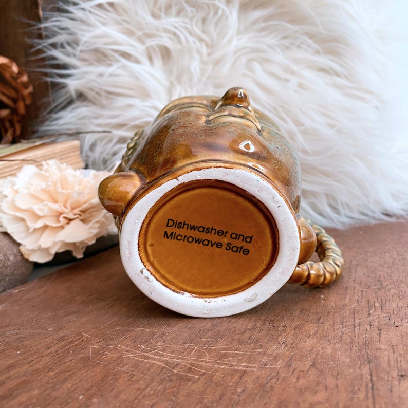 May include: A brown ceramic mug with a textured surface and a braided handle. The bottom of the mug has the text "Dishwasher and Microwave Safe" in white. The mug is on a wooden surface.