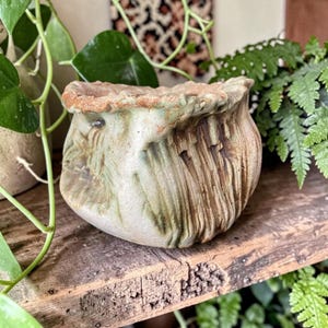 May include: A small, hand-formed ceramic planter with an irregular, wavy rim. The surface features a textured, mottled glaze in shades of sage green, cream, and earthy brown, with deep vertical grooves and a rough, unfinished clay texture at the top.