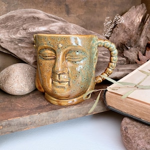 May include: A ceramic mug with a Buddha face design. The mug is primarily tan with green speckles and a textured handle. The Buddha's eyes are closed, and the mug has a serene expression. The mug is on a wooden surface.