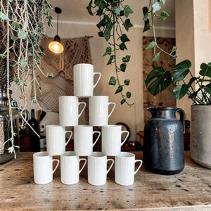 May include: A stack of nine white ceramic mugs with a vertical ribbed texture arranged in a pyramid formation, positioned next to a dark charcoal gray ceramic pitcher with a handle and a spout on a wooden surface.