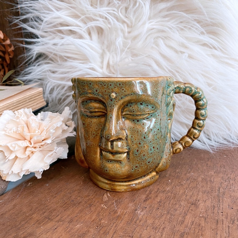 May include: A ceramic mug with a Buddha face design. The mug is primarily gold with green accents and a textured finish. The handle is a twisted rope design. The mug is on a wooden surface.