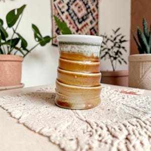Puede incluir: Un vaso cilíndrico de cerámica con un cuerpo acanalado de textura en espiral y un esmalte marrón tierra cálido. El borde superior está acabado con un esmalte blanco cremoso, liso y contrastante que gotea ligeramente por la parte superior.