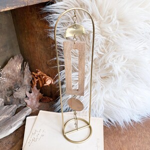 May include: A gold-toned metal arch stand featuring a hanging mobile ornament. The mobile consists of a brass semi-circle, a rectangular wooden frame, and a brass circle, all connected by small metal jump rings suspended from the top of the arch.