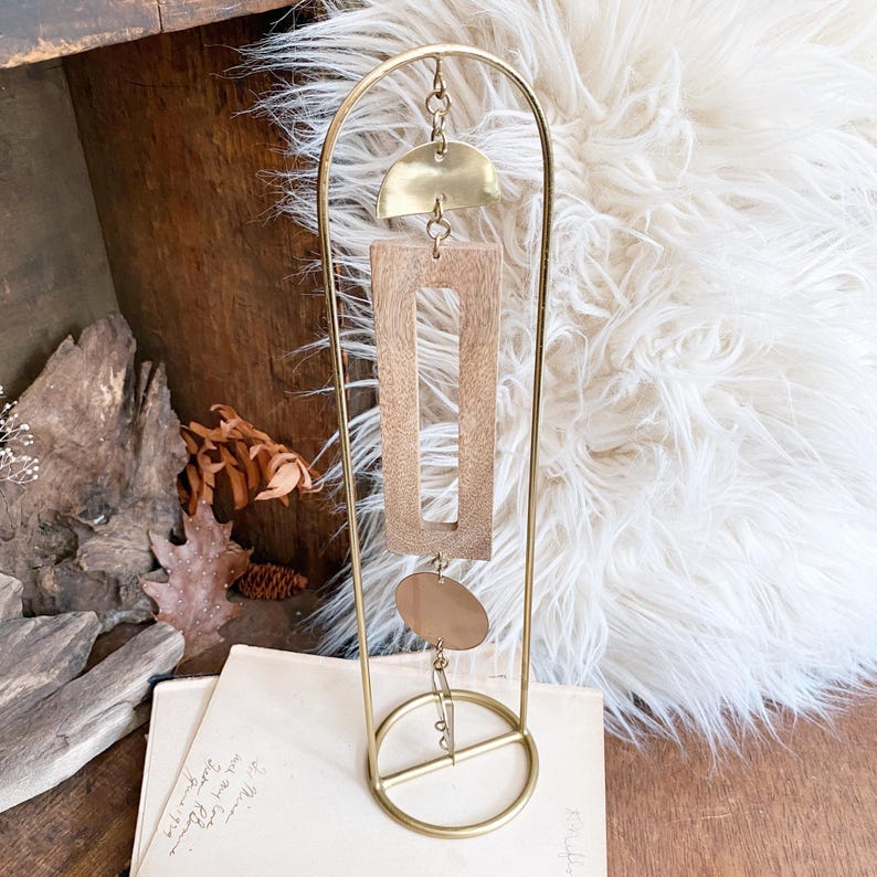 May include: A gold-toned metal arch stand featuring a hanging mobile ornament. The mobile consists of a brass semi-circle, a rectangular wooden frame, and a brass circle, all connected by small metal jump rings suspended from the top of the arch.