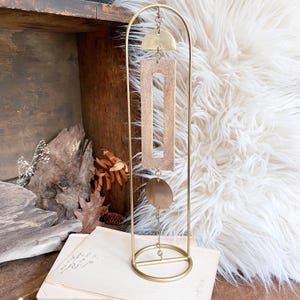 May include: A gold-toned metal arched stand featuring a hanging mobile ornament. The mobile consists of a brass semi-circle, a rectangular light-coloured wooden frame, and a circular brass disc, all connected by thin metal chains in a vertical line.