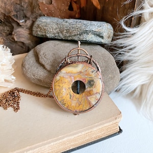 May include: A copper-toned pendant necklace featuring a circular, patterned stone with a central hole. The stone displays yellow, cream, and brown hues. The pendant is suspended from a matching copper chain, resting on a vintage book.